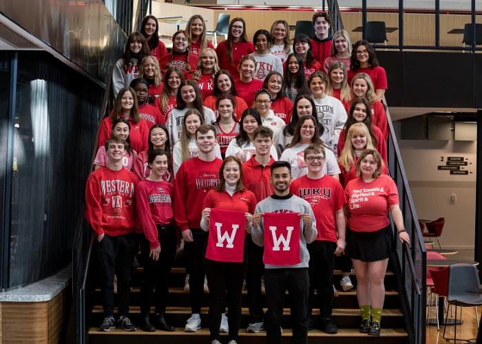 A large group of people wearing matching university-themed apparel are gathered on a staircase, smiling and holding up banners with a "W".