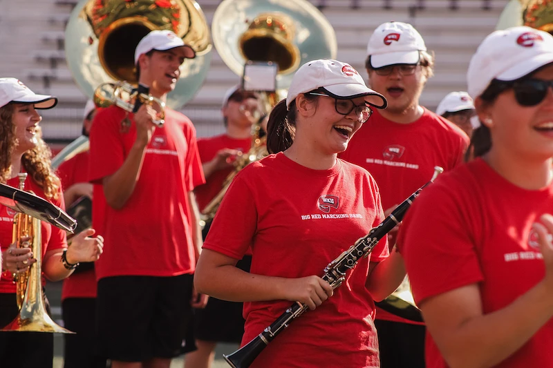 A group of marching band members in red uniforms and white hats enthusiastically play their instruments outdoors.