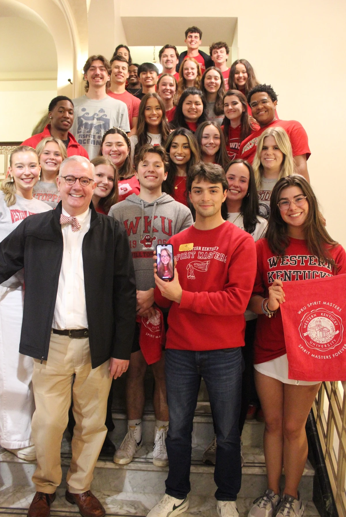 A group of smiling people, mostly wearing Western Kentucky University attire, pose on a staircase, with one person holding a phone displaying another person in a video call.