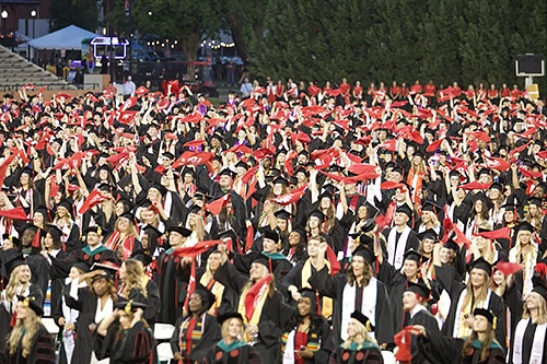 A large group of graduates in caps and gowns celebrate by waving red scarves at an outdoor commencement ceremony.
